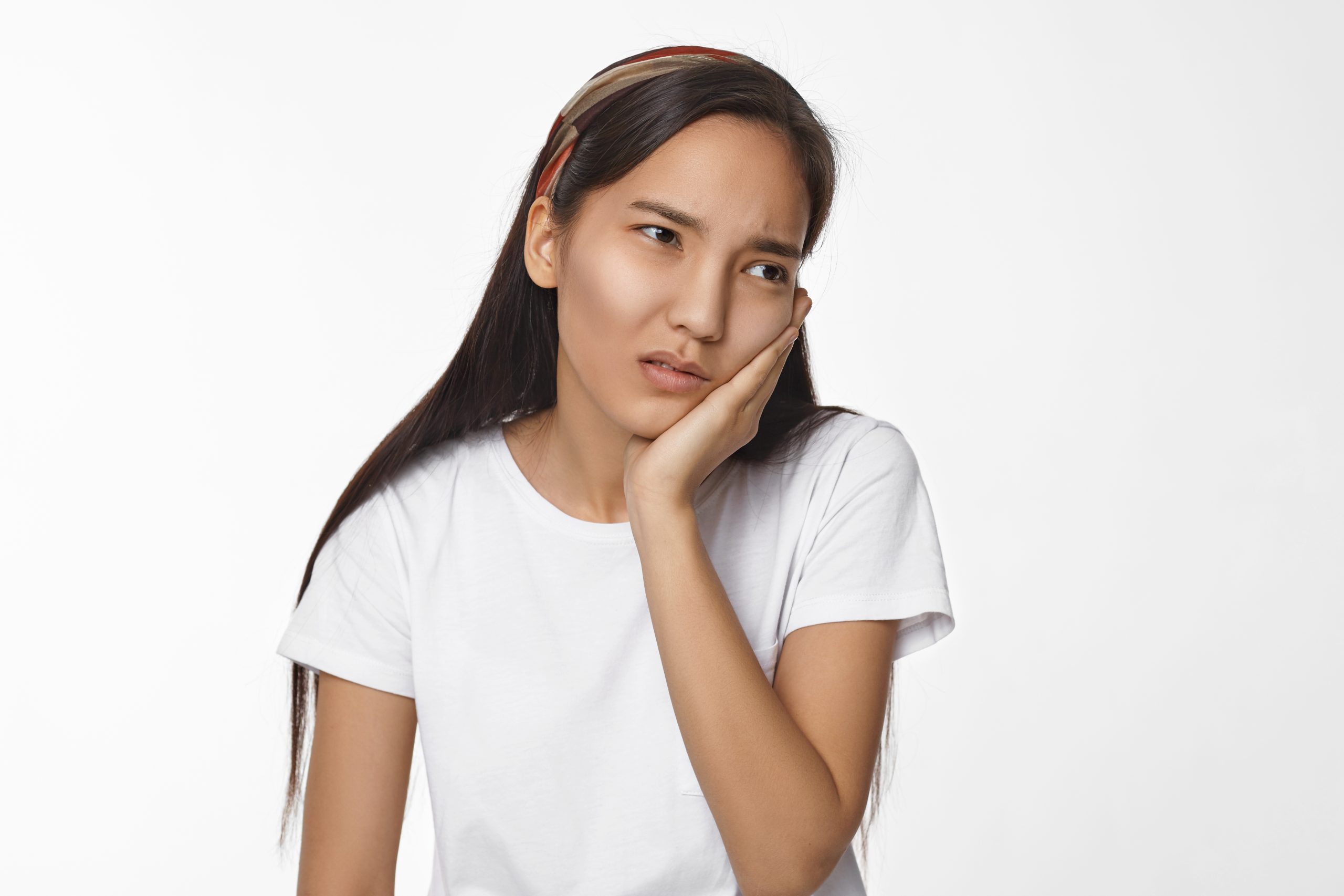 Isolated shot of dark haired frustrated young Chinese female having upset painful facial expression, holding hand on her cheek, suffering from toothache, posing isolated at white studio wall